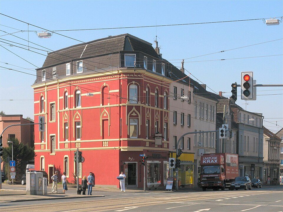 Rot gestrichenes Gründerzeit-Eckhaus mit Läden im Erdgeschoss und Wohnungen darüber an einer Straßenbahnstrecke in Bochum-Altenbochum