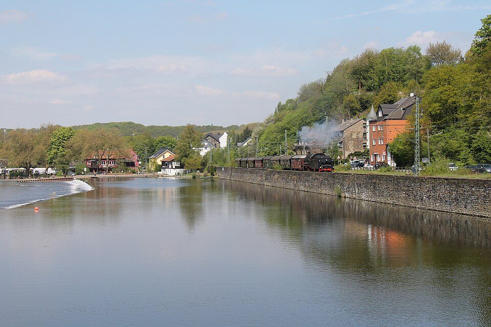 Blick über die Ruhr auf Wohnhäuser am bewaldeten Hang in Bochum-Dahlhausen, am Ufer fährt ein Museumszug der Ruhrtalbahn