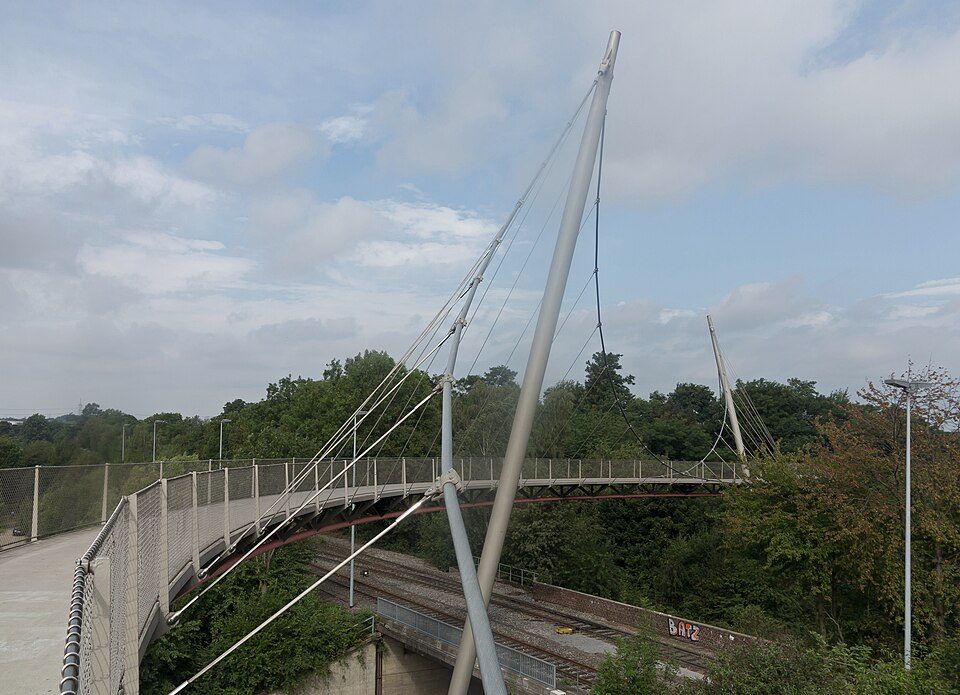 Die geschwungene Fuß- und Radwegbrücke Erzbahnschwinge mit Stahlmast und Seilen über Gleisanlagen in Bochum-Hamme