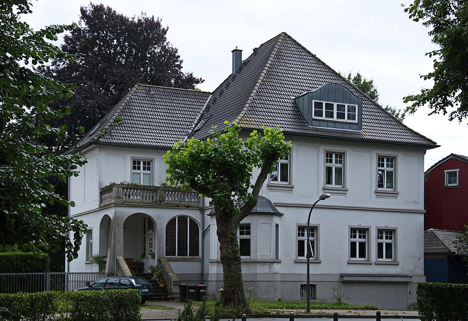 Freistehendes weißes Haus mit Walmdach, Gauben und Balkon hinter Bäumen in Bochum-Leithe, das ehemalige Arzthaus