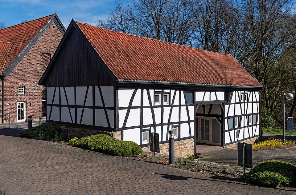 Fachwerkscheune des Beckmanns Hof mit rotem Ziegeldach und Backsteinhaus in Bochum-Querenburg, davor gepflasterter Hof und Hecken