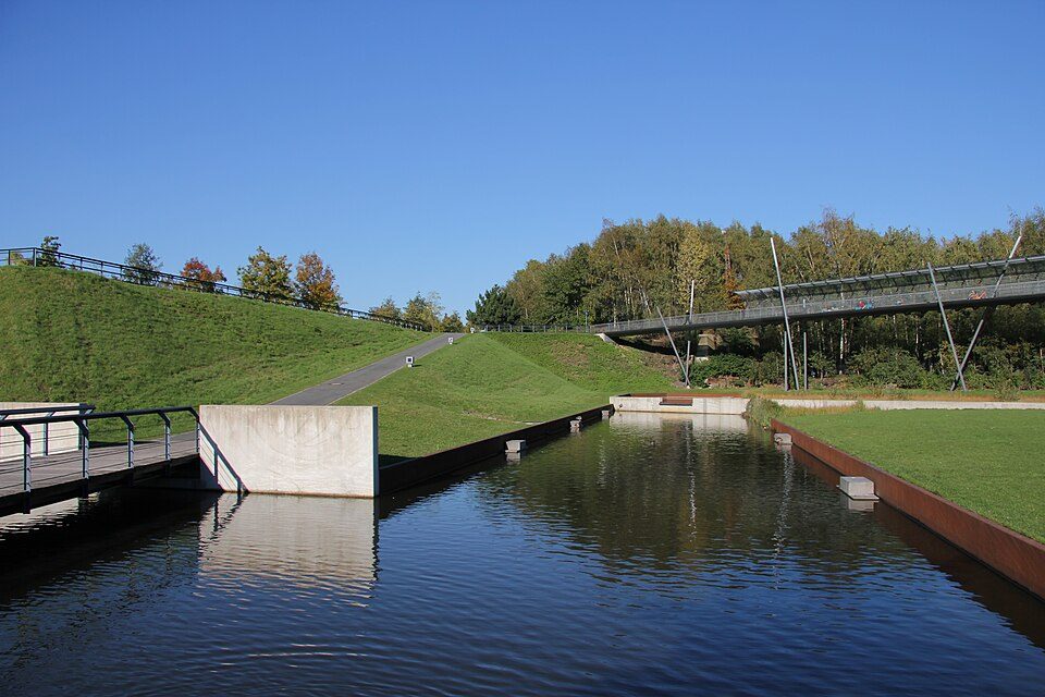 Wasserbecken im Westpark Bochum mit Steg, begrünten Hängen, einer Fußgängerbrücke und Bäumen unter blauem Himmel