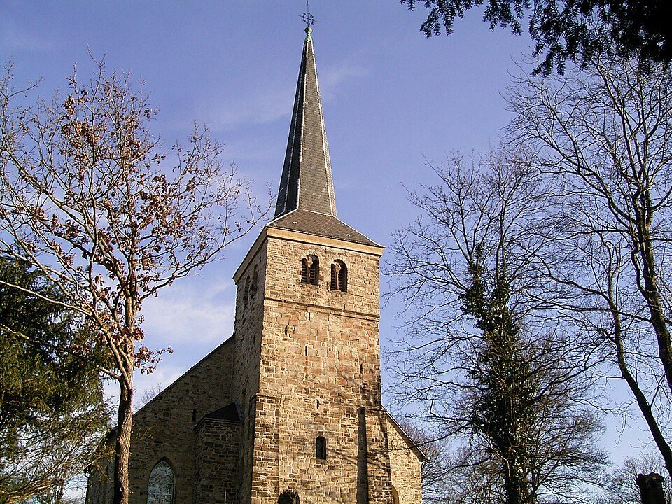 Turm der Stiepeler Dorfkirche aus Bruchstein mit spitzem Helm zwischen hohen Bäumen in Bochum-Stiepel