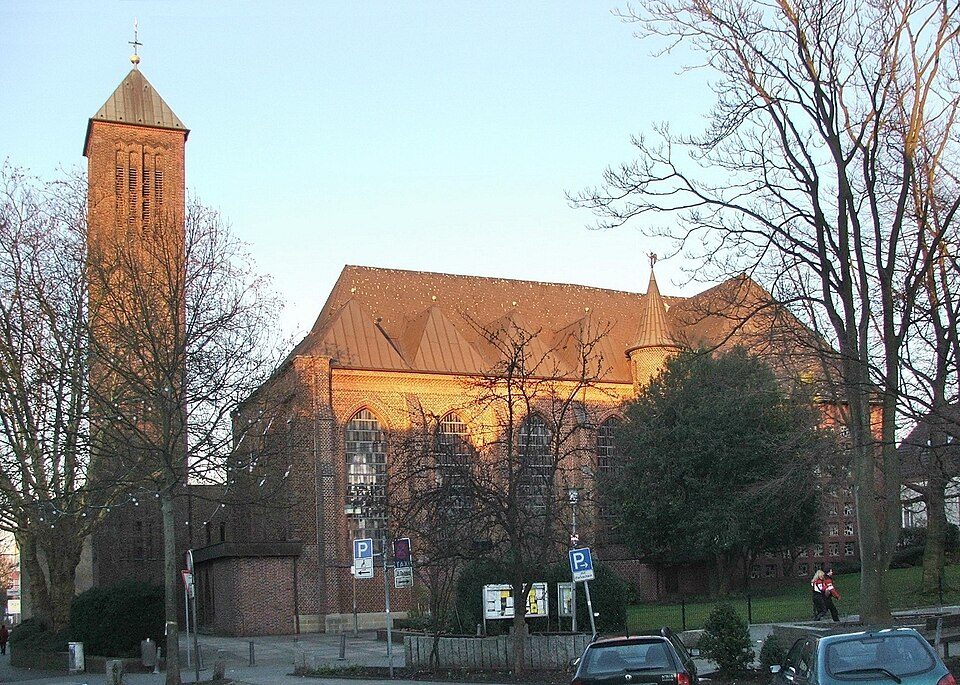 Backsteinkirche St. Johannes mit schlankem Turm im Kirchviertel von Bochum-Wiemelhausen im Abendlicht, davor Bäume und Parkplätze